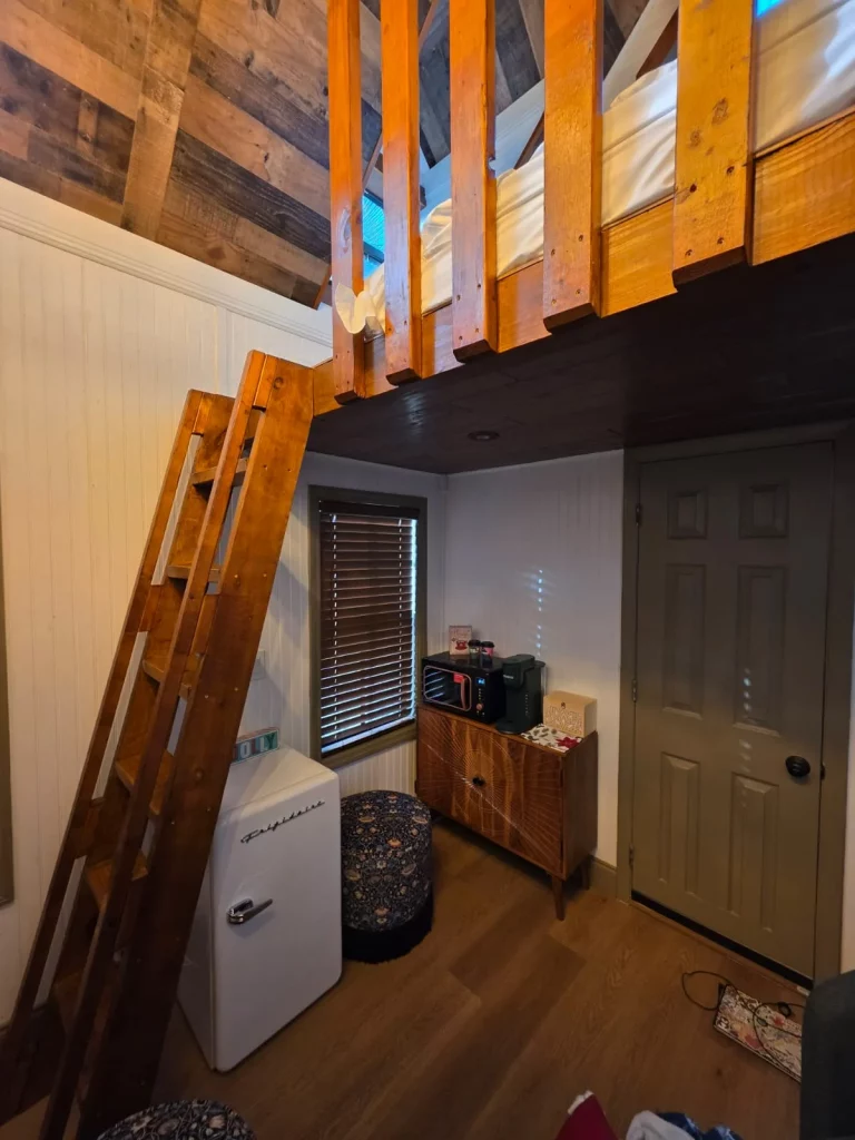 Cozy treehouse interior featuring a loft bed, wooden ladder, and mini-fridge. Perfect for treehouse getaways in Florida.